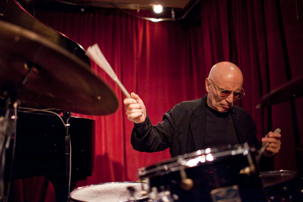 paul motian playing the drums with brushes in front of a red curtain